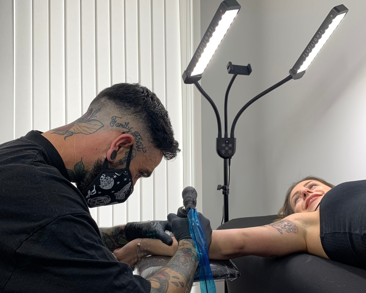 Tattoo artist working on a client with a tattoo machine, illuminated by a lamp.