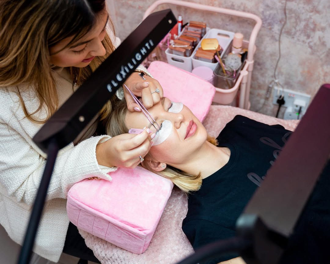 Woman getting eyelash extensions in a salon setting with a lamp and makeup products.